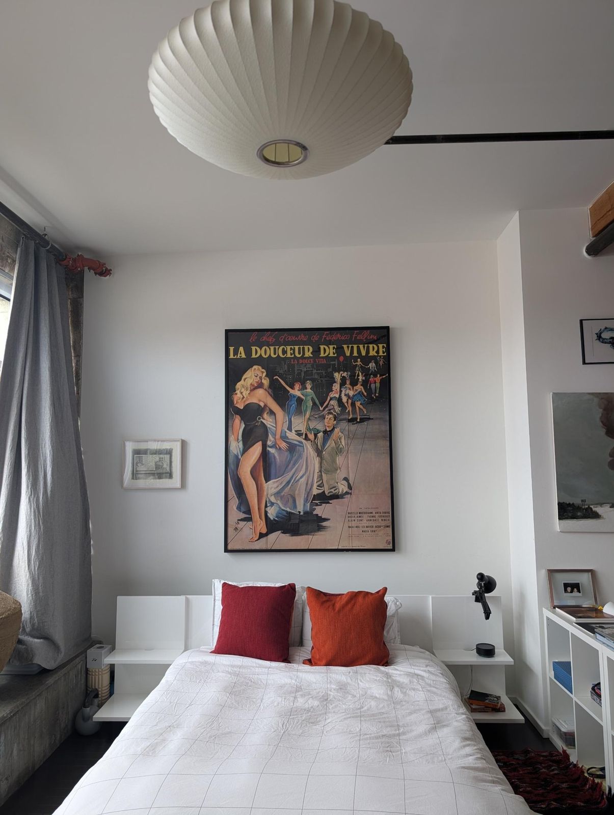A white bed with red pillows under a paper bubble pendant, with a vintage film poster above.