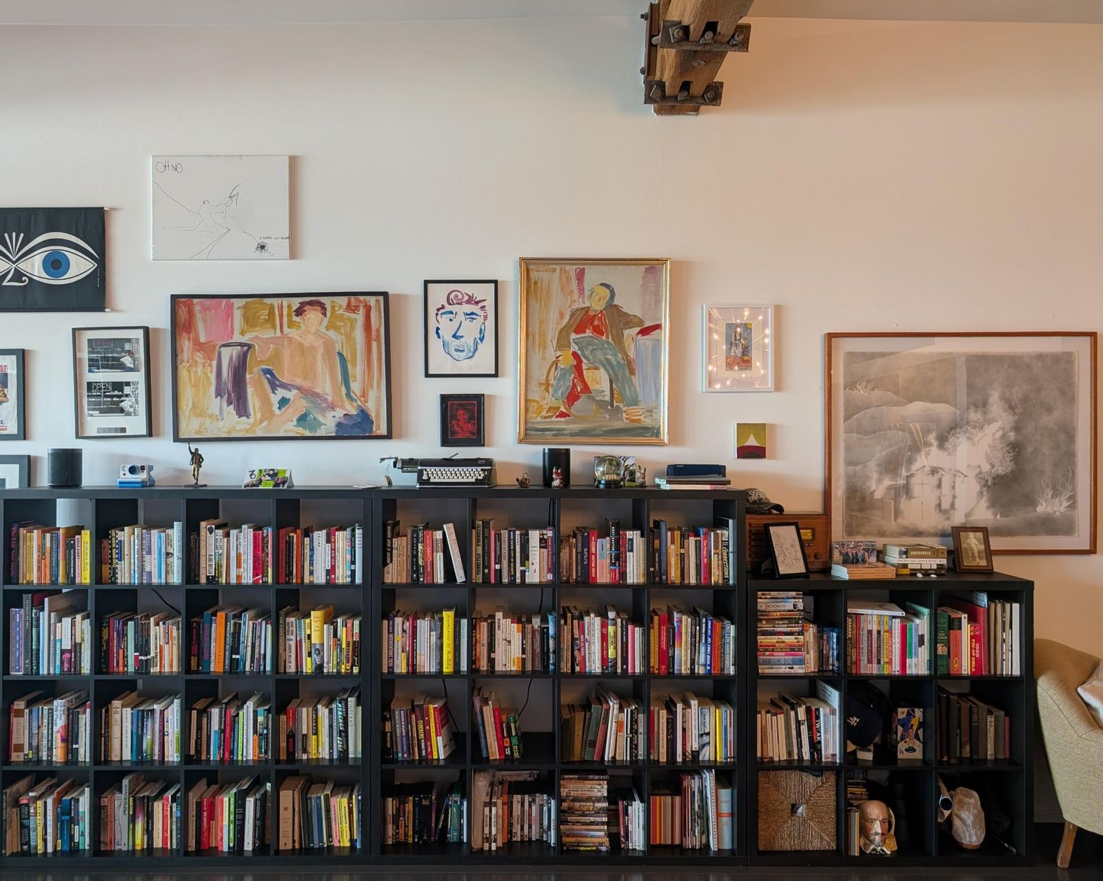Paintings, prints and drawings hung salon-style above a long black bookcase.