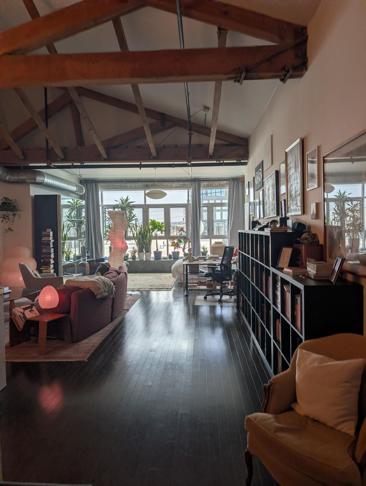 Long view of a loft great room under exposed timber trusses, toward a wall of windows lined with plants.