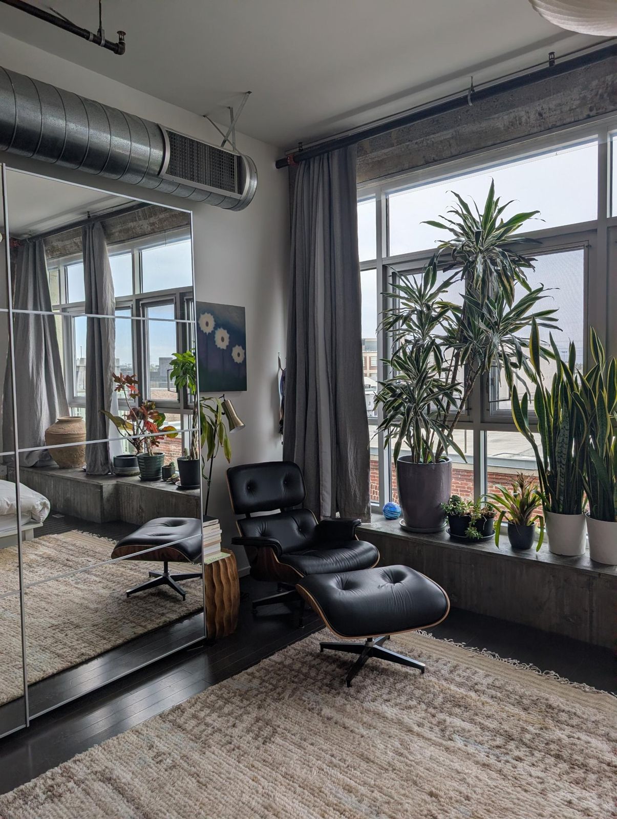 A black leather Eames lounge chair on a deep wool rug beside tall windows and potted plants.