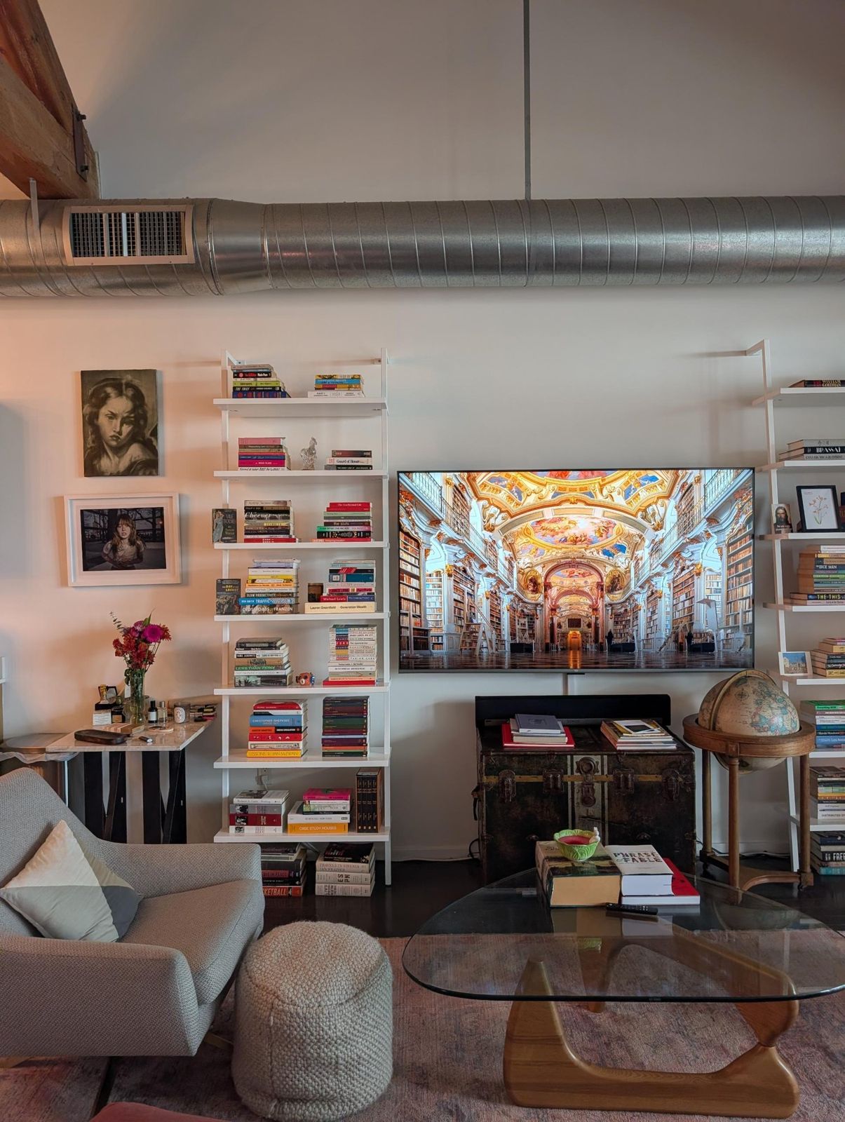 White ladder shelves of stacked books flanking a wall-mounted screen, with two portraits hung to the left.