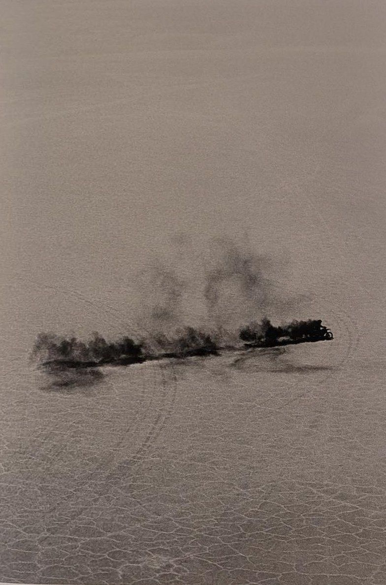 Black-and-white photograph of a dark plume of dust trailing across a pale salt flat.